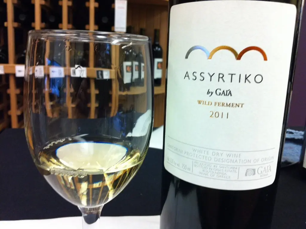 A glass of pale Assyrtiko white wine beside a bottle in a wine shop
