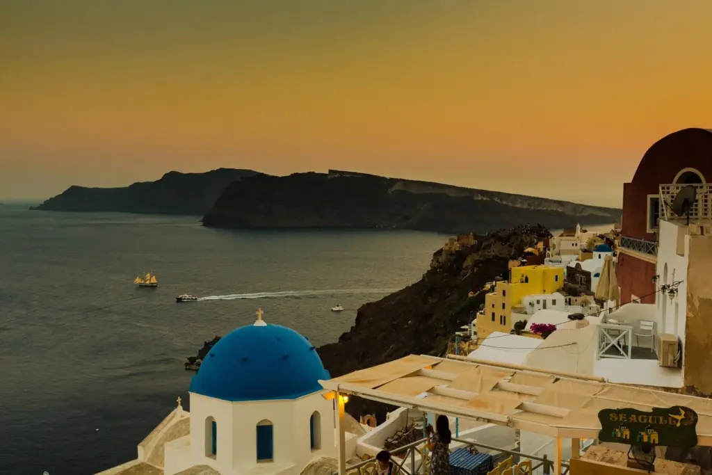A blue-domed church and cliffside houses above the caldera at sunset, with Thirasia on the horizon