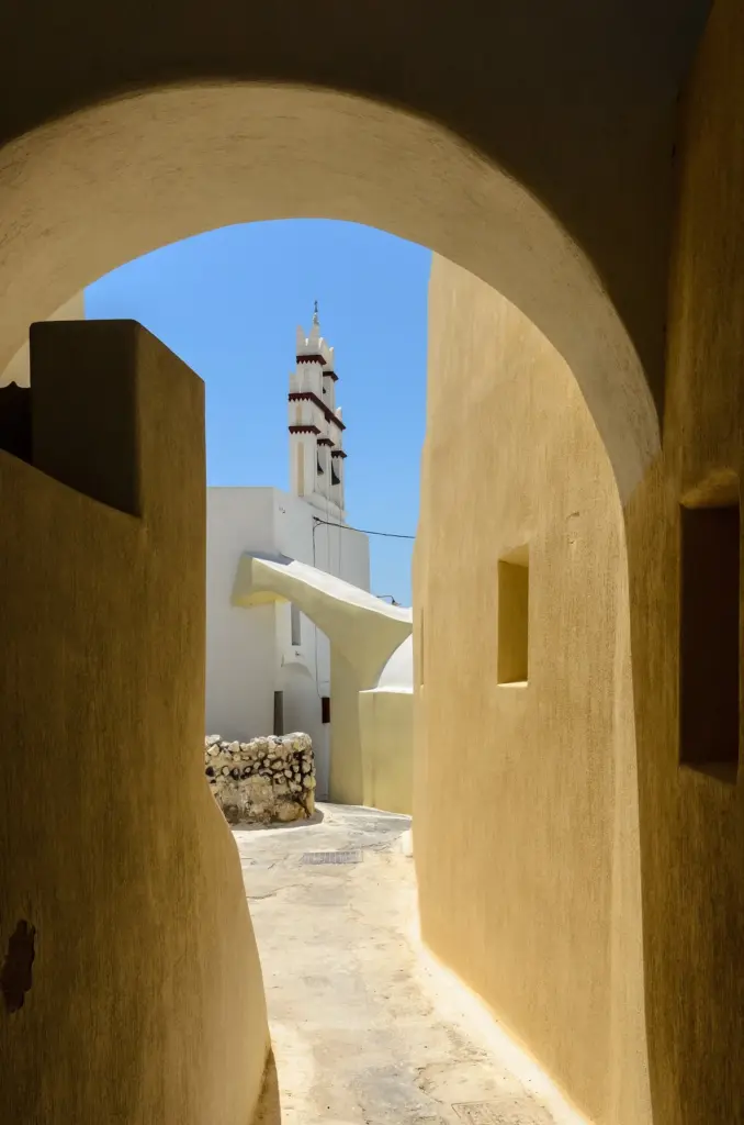 A narrow sunlit lane under a stone arch in Emporio's old quarter, with a white bell tower beyond