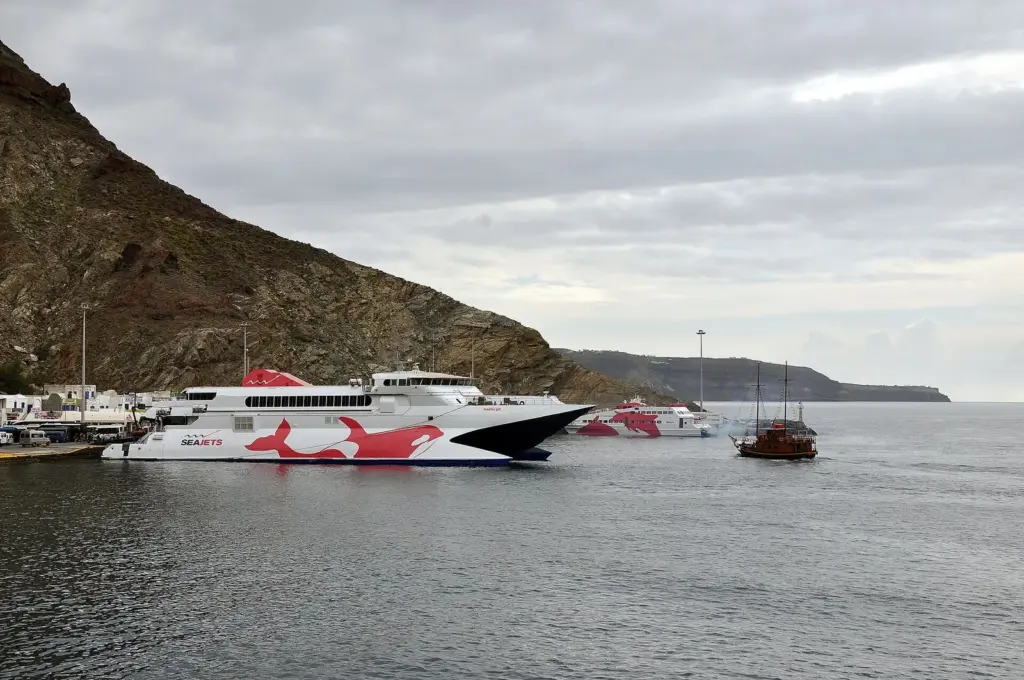 A white and red Seajets high-speed catamaran at Athinios port under the cliffs, with a wooden schooner nearby