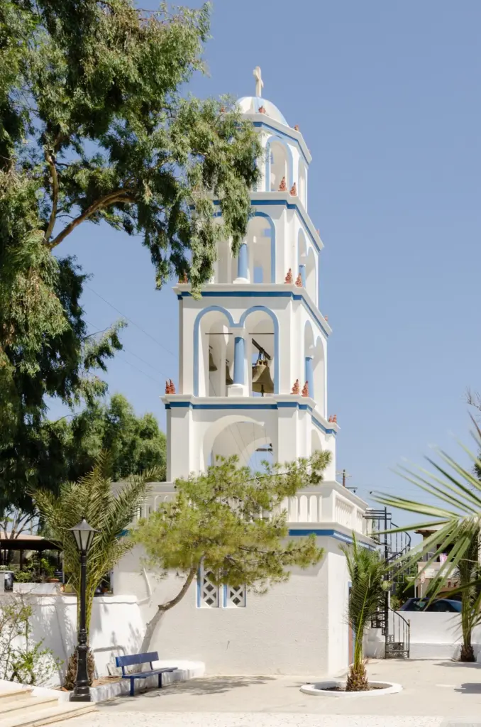 A white and blue church bell tower among trees and palms in Kamari