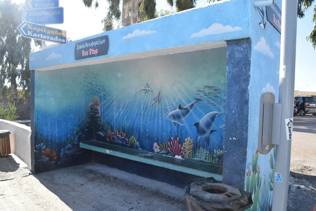 A bus stop shelter painted with a colorful sea mural and signs pointing to the airport and Karterados