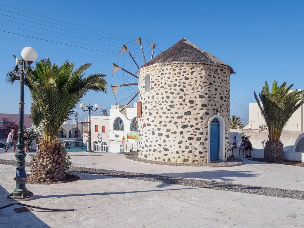 A round stone windmill with a thatched roof on the main square of Karterados, framed by palm trees