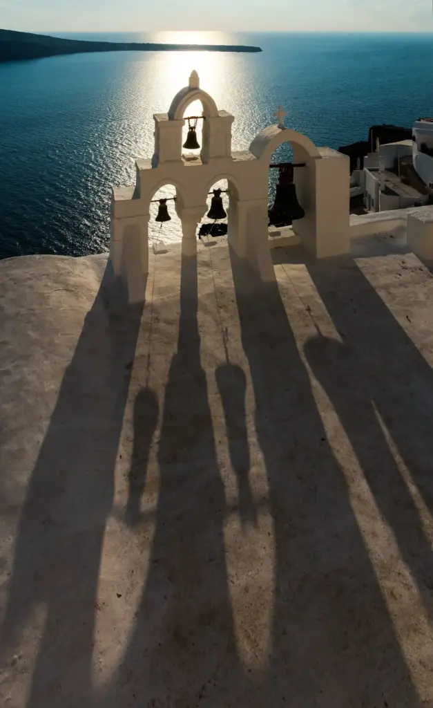A white bell tower with four bells casting long shadows on a rooftop above the sea in Oia