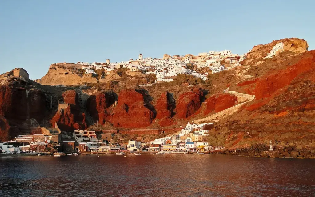 The red cliffs of Oia rising above a small harbor, with the village along the clifftop and a stepped path zigzagging down