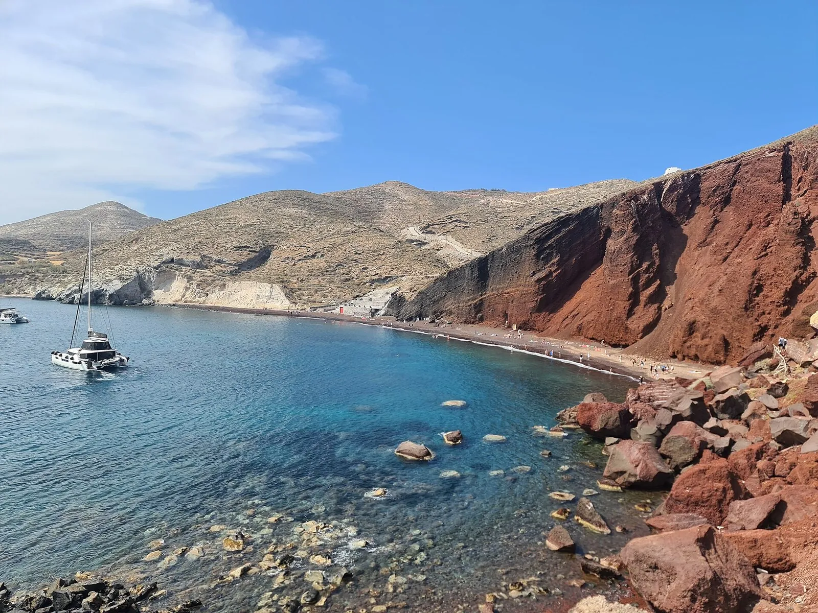 Santorini beaches: the rust-red lava cliffs and turquoise water of Red Beach with a catamaran at anchor