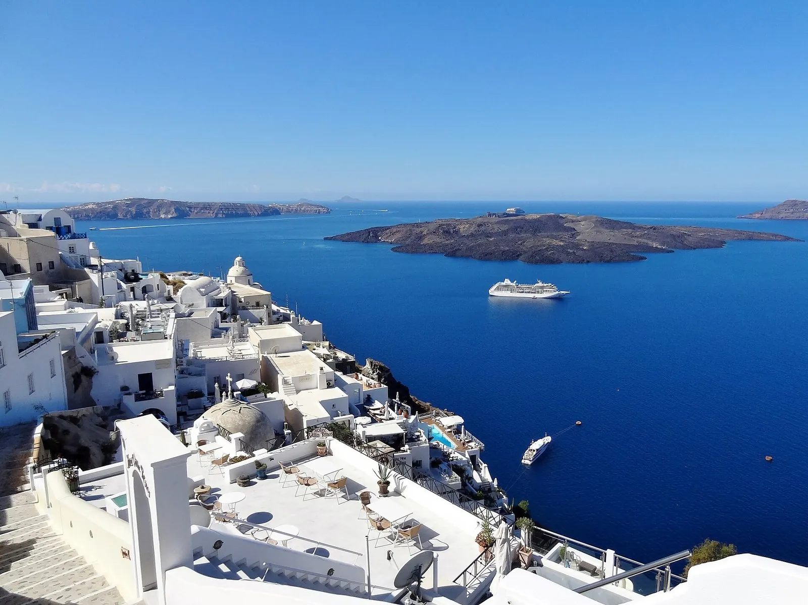 Santorini hotels with plunge pools and terraces stepping down the caldera cliff in Fira, with the Nea Kameni volcano and a cruise ship in the bay