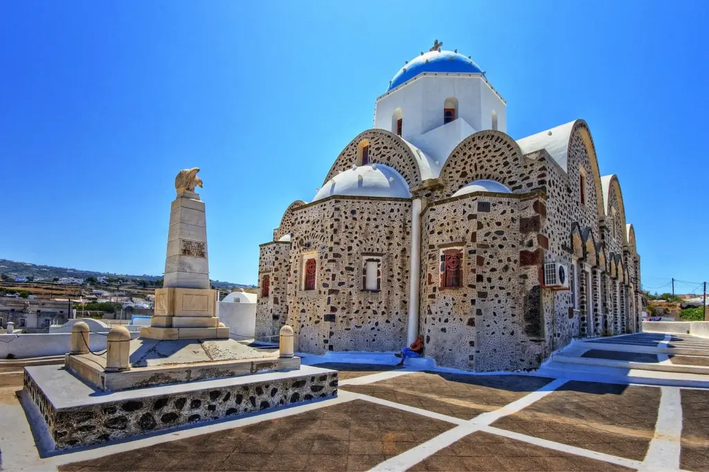 A stone church with arched roofs and a blue dome beside a monument on a square in Vothonas