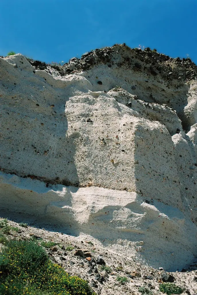 A tall cliff of pale layered pumice and ash under a blue sky, with plants at its foot