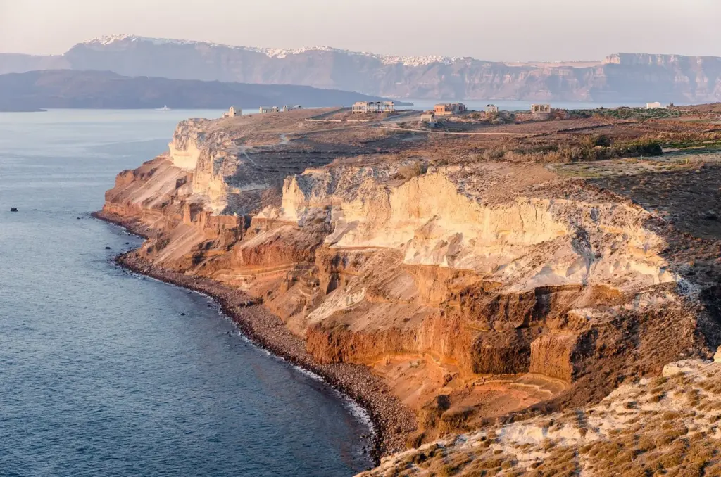 Pale layered cliffs at Cape Akrotiri glowing in evening sun, with the caldera rim and its villages in the distance