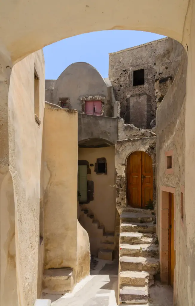 Narrow cream-colored lane under a stone arch, with steps, small domes and a wooden door in Emporio