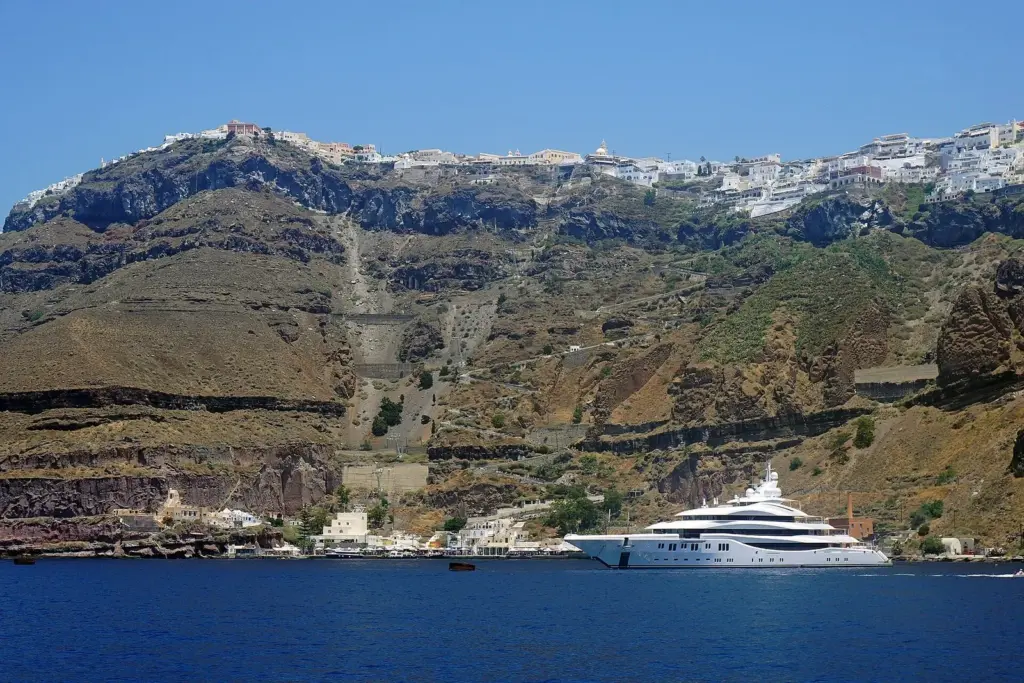 Fira's white buildings on the cliff top above the cable car line and the zigzag steps down to the small port, with a large yacht offshore