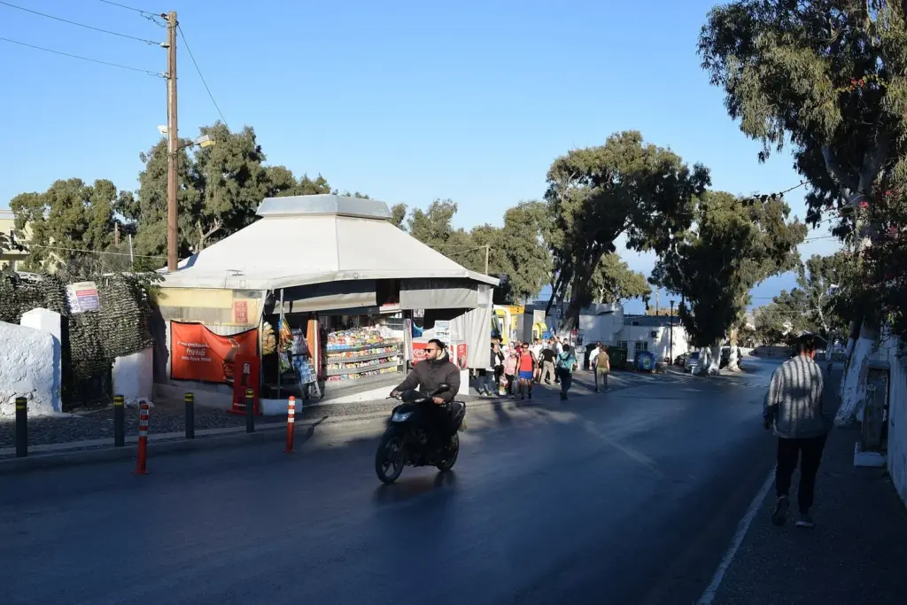 A kiosk and shaded waiting area at Fira's main bus station, with a scooter rider on the road
