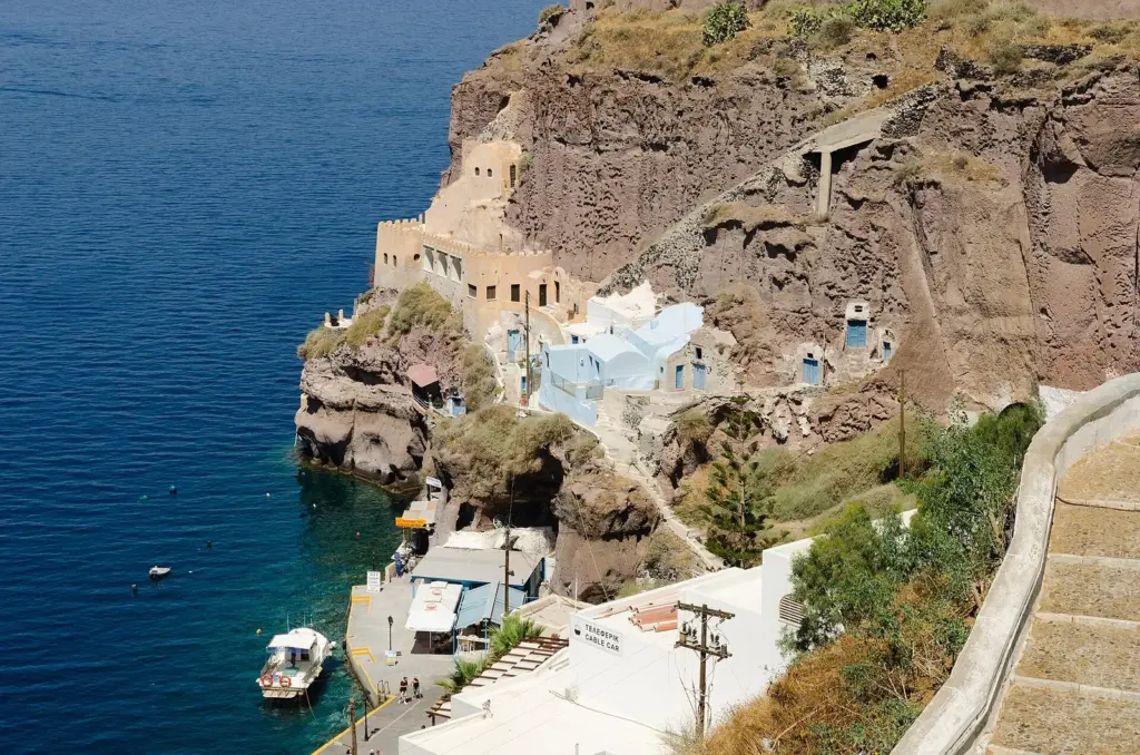 Cave houses and whitewashed buildings cut into a brown cliff above a small harbor with a moored boat