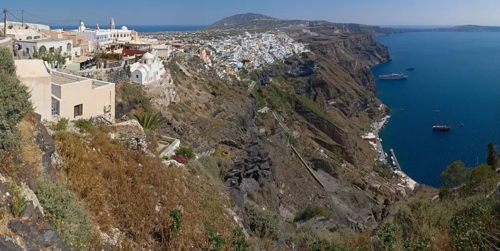 The road and steps winding down a dry cliff from Fira's white buildings to the sea, with boats in the bay