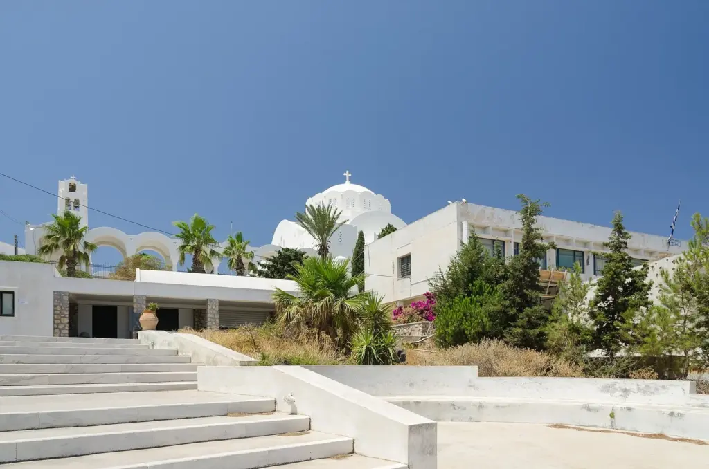 White Orthodox cathedral dome and bell arches behind a low museum building, palm trees and wide steps in Fira