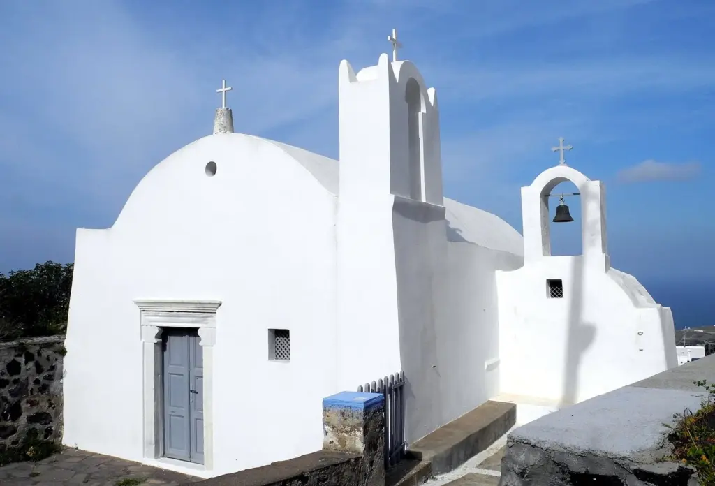A whitewashed church with a bell gable and a stone-framed door in Firostefani under a blue sky