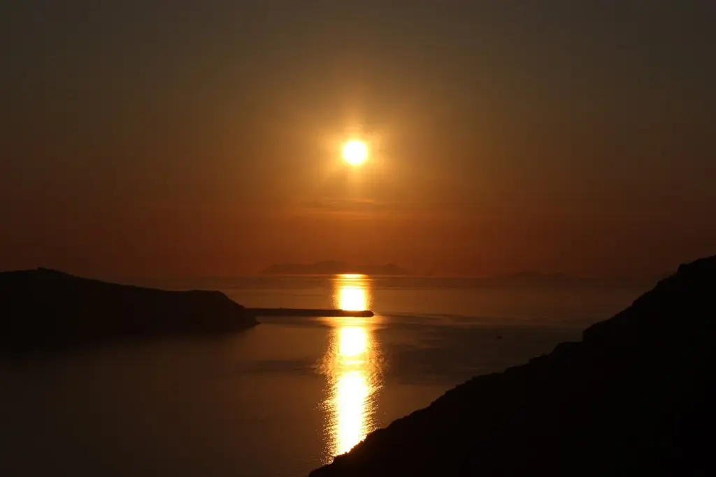 The sun setting over the caldera, reflected in the sea between dark headlands, seen from Firostefani