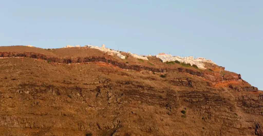 The red and brown layered cliffs below Imerovigli lit by evening sun, with white buildings along the top