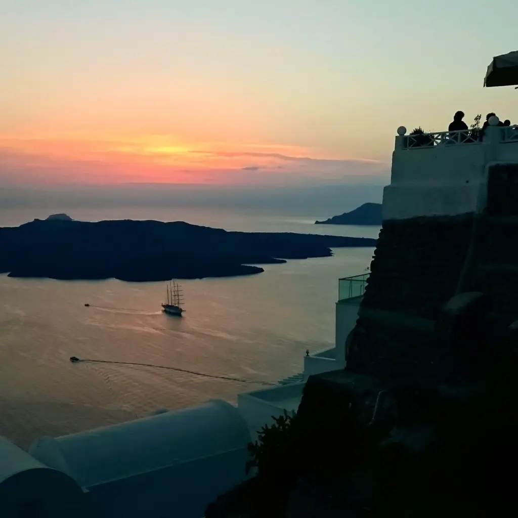 A tall-masted sailing ship on the calm caldera at sunset, with dark islands and cliff-top terraces in silhouette