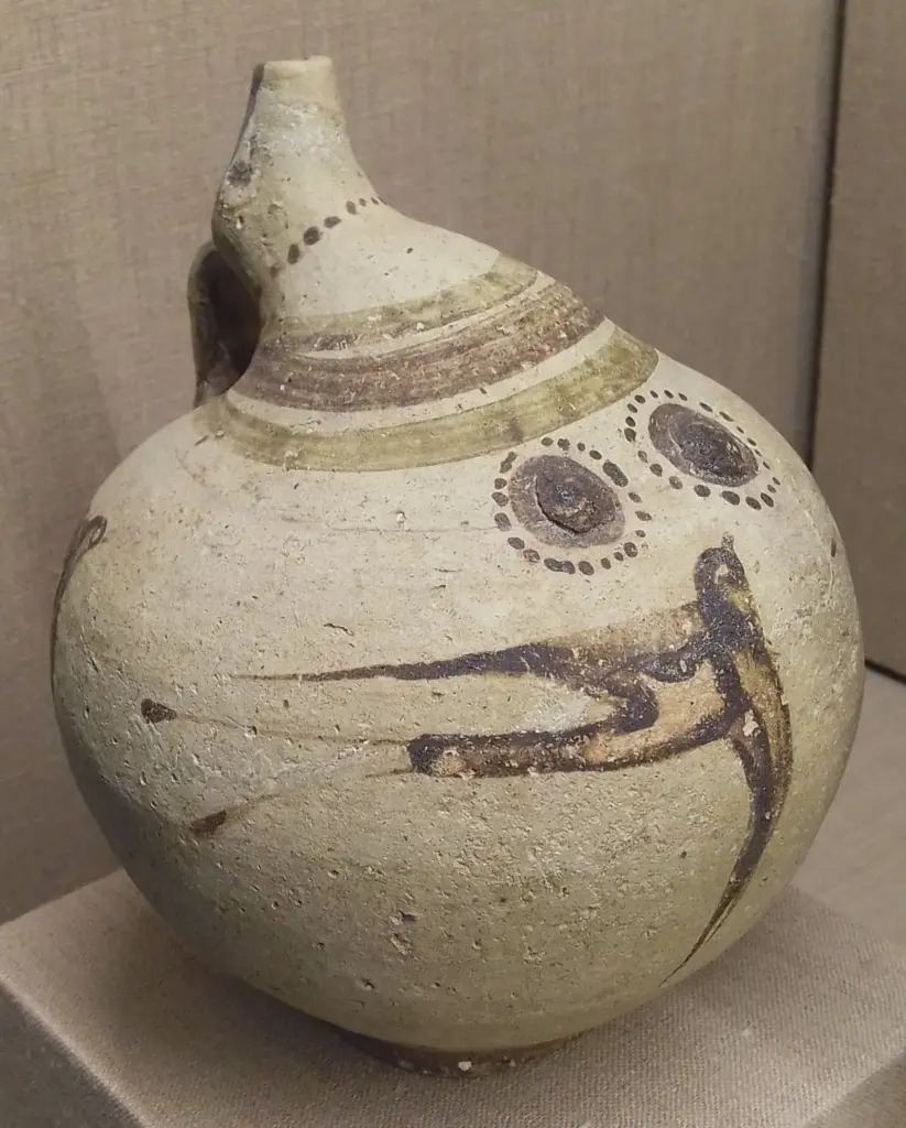 Pale clay jug with a long spout and a dark swallow painted on its body, in a museum case