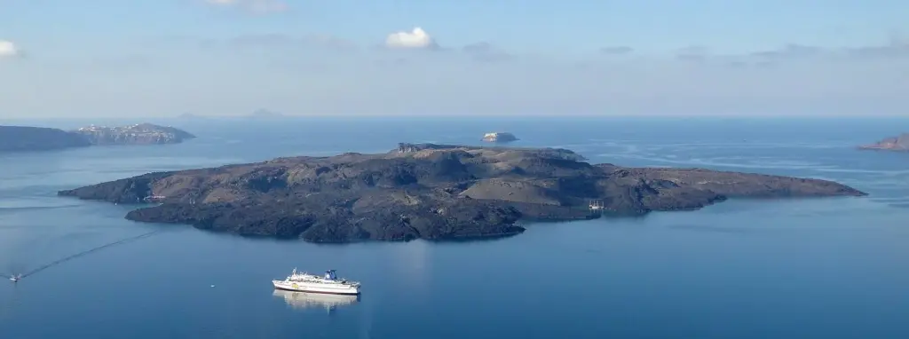 The dark lava island of Nea Kameni in the middle of the blue caldera, seen from the cliffs above, with a ship nearby