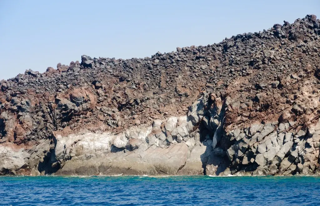 Jagged dark lava blocks above a band of pale rock at the waterline of Nea Kameni