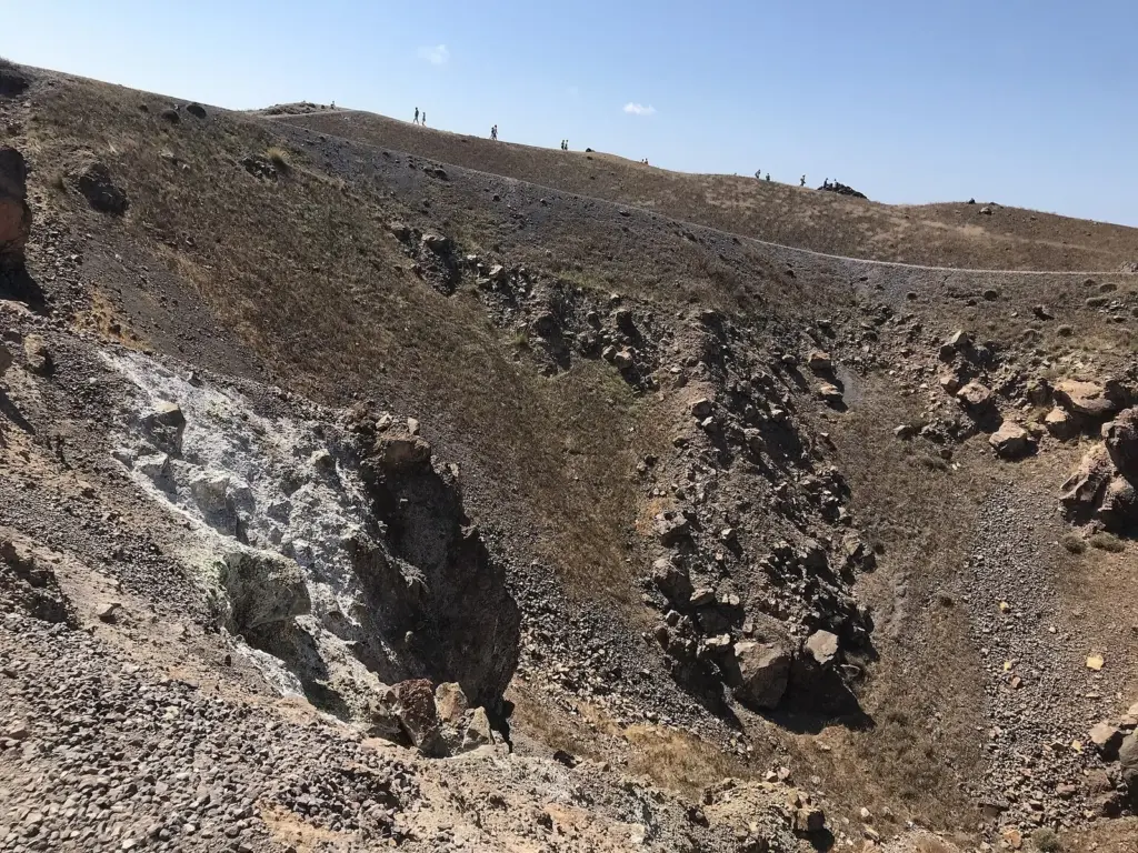 A barren crater of brown cinders and boulders on Nea Kameni, with hikers on a path along its rim