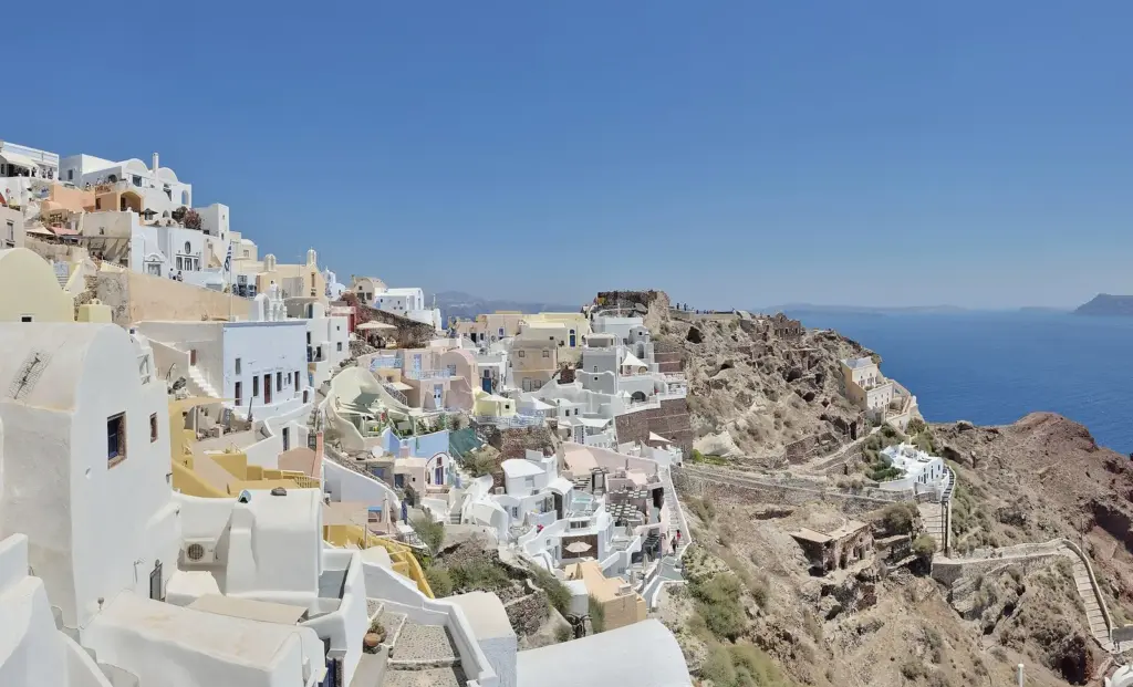 White and pastel houses of Oia spread along the top of the caldera cliff above a dry slope and the sea