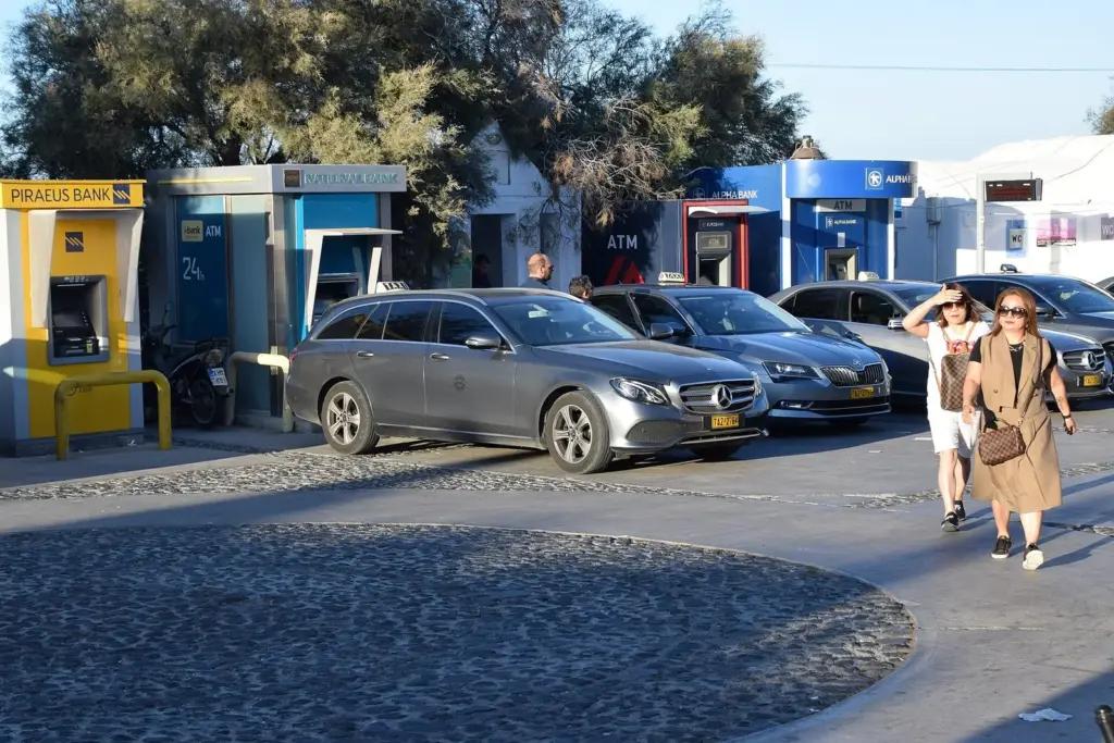 Taxis and cars waiting by a row of bank ATMs on a square in Oia, with people walking past