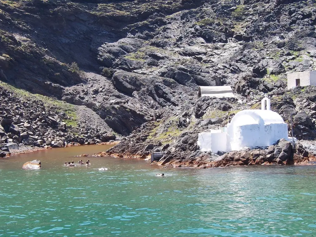 Swimmers in the shallow orange-tinted water of a rocky inlet beside a small white chapel