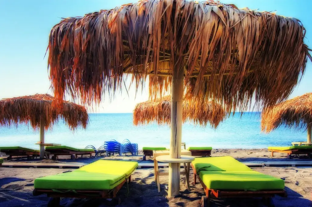 Thatched umbrellas and green sunbeds on a dark-sand beach facing a calm blue sea