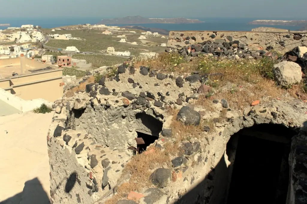 Crumbling dark volcanic-stone walls of a castle above village rooftops, with islands in the caldera beyond