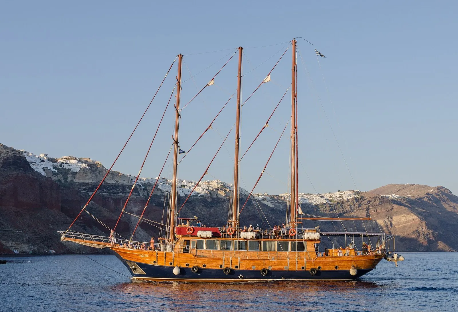 A three-masted wooden tour boat sailing in the caldera below the cliffs of Oia on a Santorini boat tour
