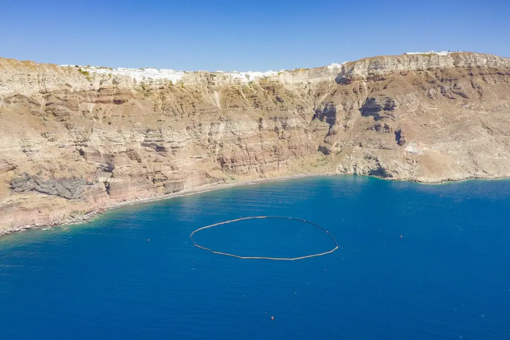 Banded caldera cliffs in shades of red, brown and cream rising from deep blue water, with white buildings along the top