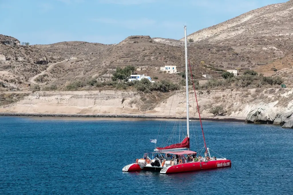 A red catamaran with its sail lowered anchored in a calm blue bay below dry hills