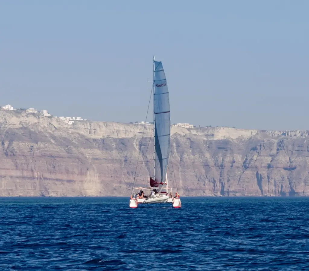 A white catamaran under sail on deep blue water in front of the tall caldera cliffs