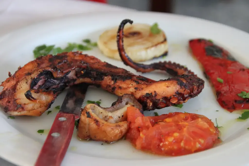 Grilled octopus tentacle with charred onion, red pepper and tomato on a white plate