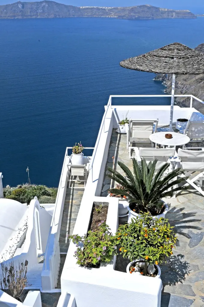 A whitewashed hotel terrace with a table, potted plants and a thatched umbrella above the blue caldera