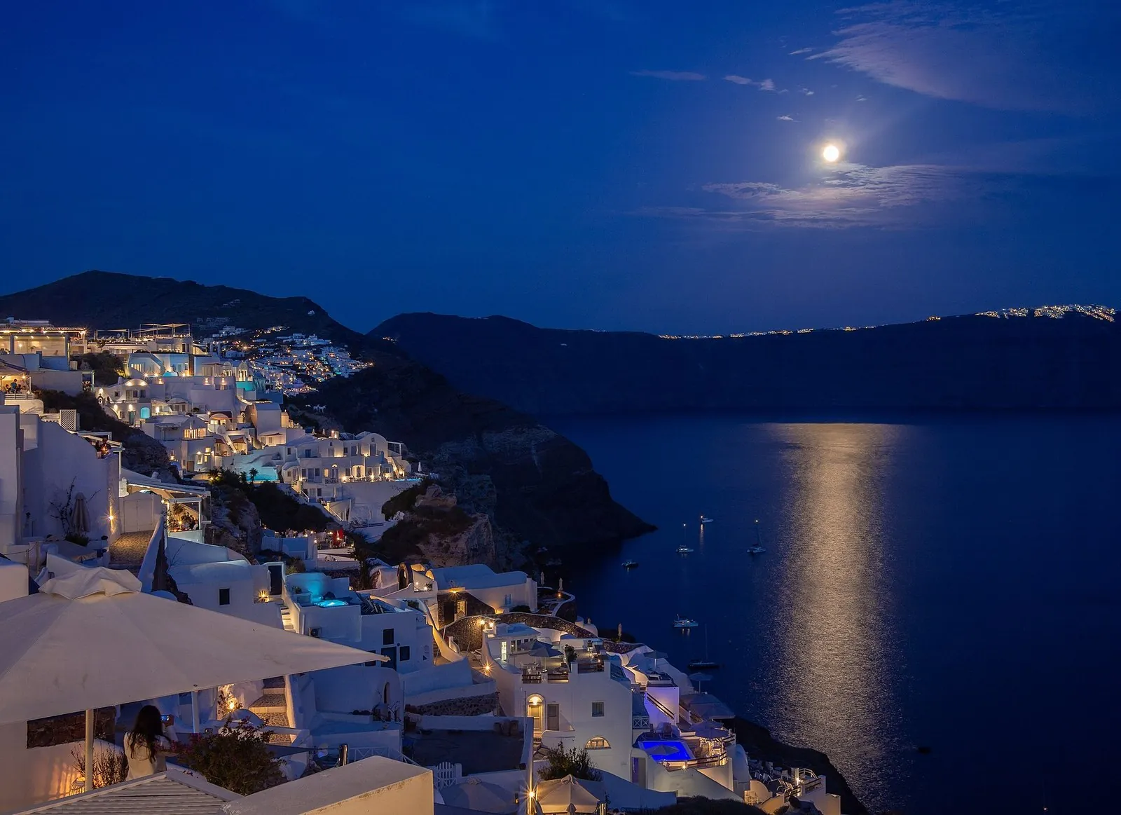 Oia's white houses lit up at dusk under a rising moon over the caldera, the kind of evening a Santorini itinerary is built around