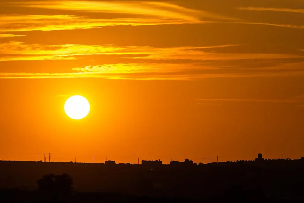 The sun rising in an orange sky above the dark silhouette of a hill and village on Santorini