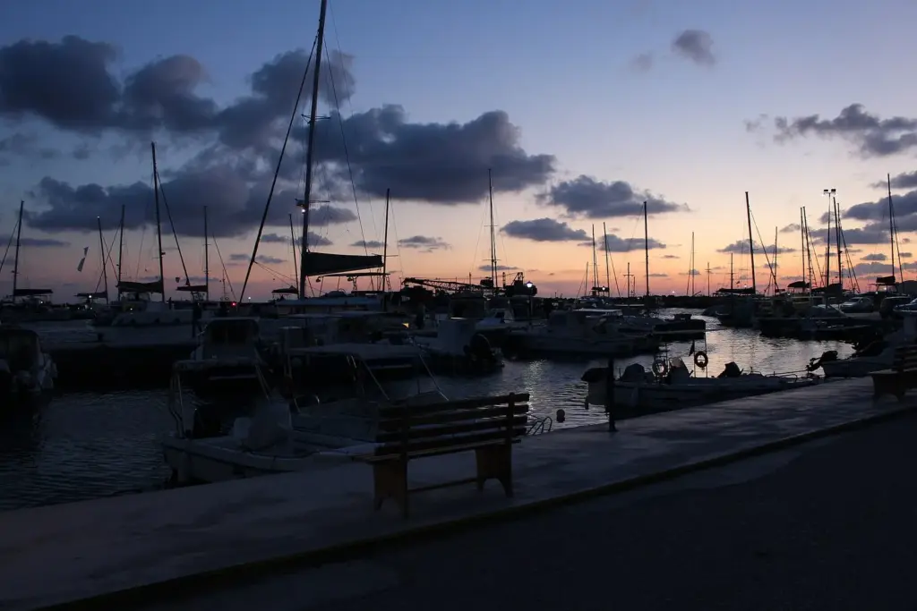 Sailboat masts silhouetted against a dusky sky at Vlychada marina, with benches on the quay